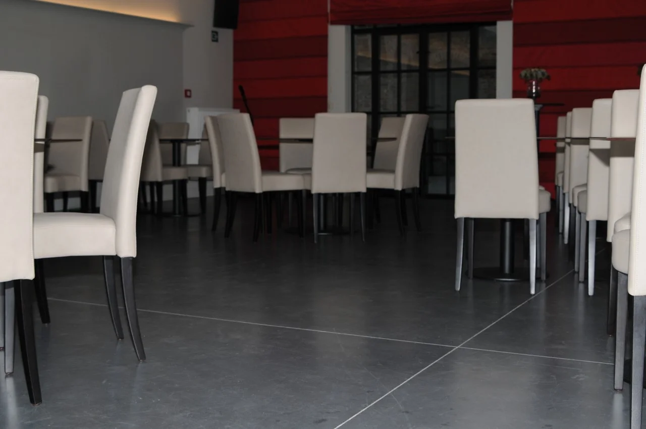 Donkere gevlinderde betonvloer met zaaglijnen tussen horecatafels en witte stoelen